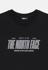 Melnas kokvilnas T-krekls ar apaļu kakla izgriezumu, uz kura ir drosmi balta teksts "THE NORTH FACE" un "NEVER STOP EXPLORING" kopā ar ģeogrāfiskiem koordinātēm.