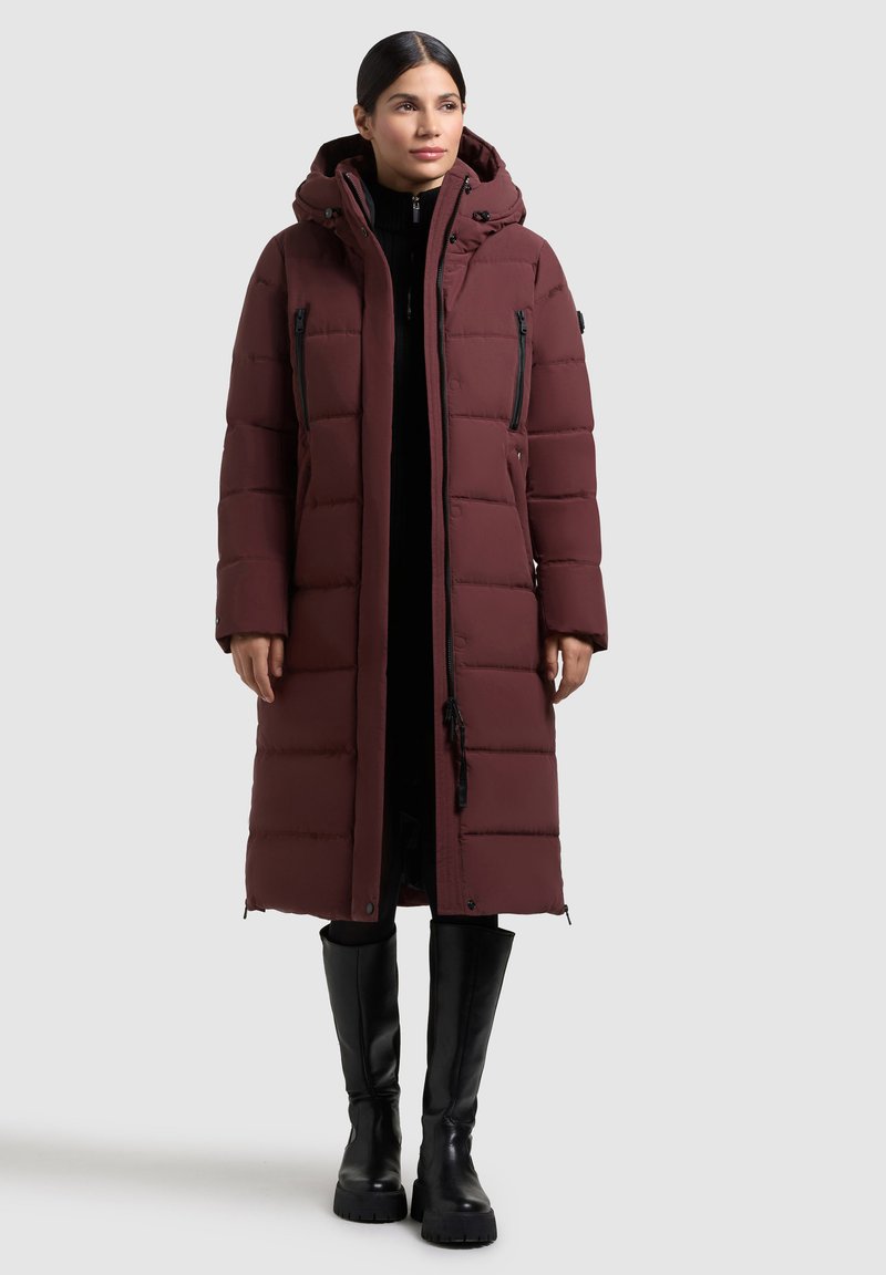Abrigo acolchado burdeos con capucha, que presenta secciones acolchadas y cremalleras, combinado con botas negras hasta la rodilla para mayor calidez y cobertura.