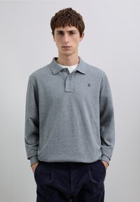 Polo à manches longues gris avec un col à revers et une patte de boutonnage à bouton unique. Comprend un petit logo sombre sur le côté gauche de la poitrine.