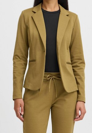 Blazer vert olive en tissu doux avec une coupe ajustée, des revers crantés et deux poches avant. Porté sur un haut noir.