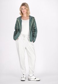 Jachetă impermeabilă verde cu fermoar galben, tricou alb, pantaloni de trening gri deschis și adidași alb-negru. Look casual de îmbrăcăminte sport.