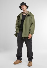 Lettvektig olivengrønn jakke med hette, svart skjorte, svarte cargo-bukser og brune støvler, med elastiske mansjetter og sidelommer.