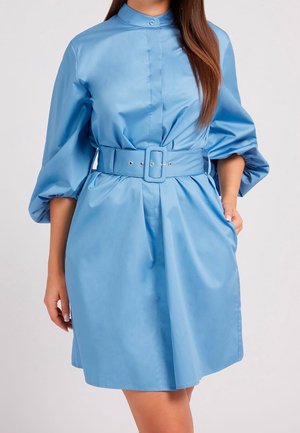 Frau trägt ein hellblaues Kleid mit Gürtel, Puffärmeln und hohem Kragen und steht mit einer Hand in der Tasche.