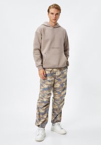 Sudadera beige con bolsillo frontal y texto impreso. Pantalones con patrón de camuflaje en beige, azul y marrón, combinados con zapatillas blancas.