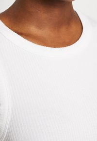 Top bianco in cotone a coste con una texture morbida e una scollatura rotonda, che mostra i dettagli ravvicinati del tessuto e delle cuciture.