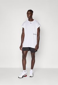 T-shirt sans manches blanche avec le logo PUMA noir, associée à un short noir et des baskets blanches avec des accents rouges. Vêtements de sport simples et décontractés.