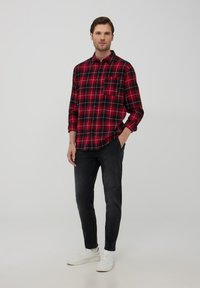 Camicia a quadri rossa e nera con maniche lunghe, abbinata a jeans scuri e sneakers bianche, in piedi su uno sfondo neutro.