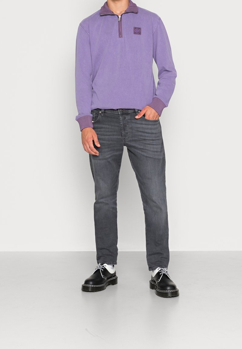 Personne portant un pull violet à fermeture éclair, un jean gris foncé, des chaussettes blanches et des chaussures en cuir noires à lacets, debout devant un fond uni.