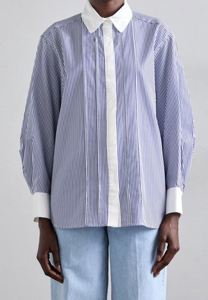 Osoba ubrana w niebiesko-białą bluzkę w pionowe paski z białym kołnierzykiem i mankietami, w połączeniu z jasnoniebieskimi dżinsami.