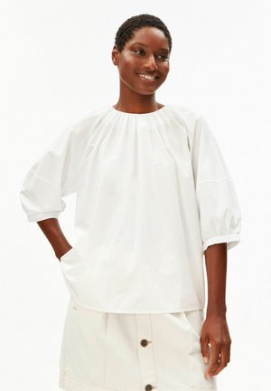 Glimlachende vrouw met kort haar, die een loszittende witte blouse met pofmouwen en een lichtgekleurde rok met knopen draagt.