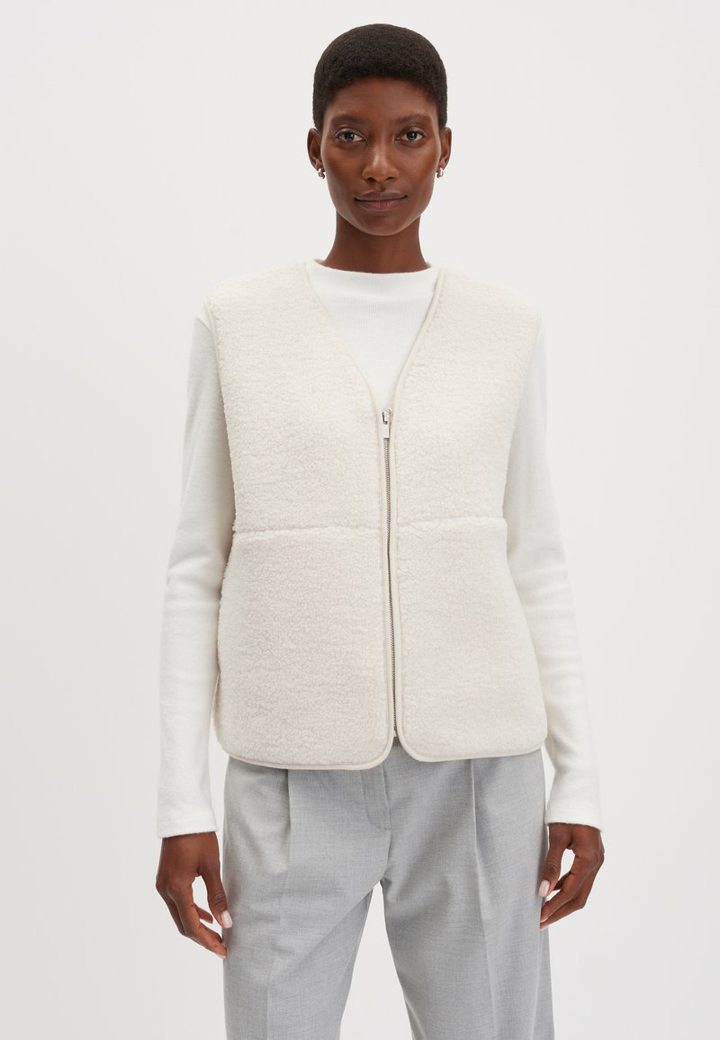 Fleeceweste in Off-White, mit durchgehendem Reißverschluss, ärmellosem Design und glatter Textur. Trägt man über einem weißen Rollkragenpullover und grauen Hosen.