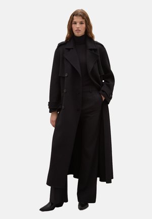 Cappotto lungo nero con design doppiopetto, ampi revers e tasche laterali, abbinato a un dolcevita nero e pantaloni sartoriali.