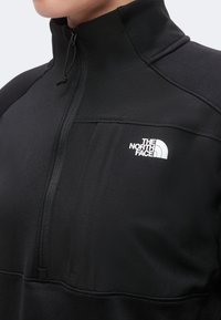 Gros plan d'une personne portant une veste noire zippée avec un col montant et le logo The North Face sur la poitrine.