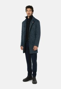 Wollen blend overcoat in donkerblauw met een gestructureerde afwerking, voorzien van een hoge kraag en knoopsluiting. Gekleed met donkere broek en enkellaarzen.