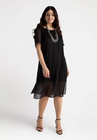 Zwarte jurk met een vloeiende chiffon onderlaag, korte mouwen en een vierkante halslijn. Aangevuld met een multi-kleurige kralenketting.