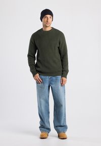 Hombre con gorro negro, suéter acanalado verde oliva, jeans de pierna ancha azul claro y botas de trabajo color beige, de pie contra un fondo sencillo.