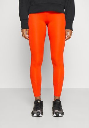 Person trägt leuchtend orangefarbene Leggings, schwarze Sportschuhe und ein schwarzes langärmeliges Oberteil und steht vor einem einfarbigen Hintergrund.