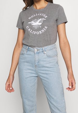 T-shirt gris en coton avec un motif blanc "Hollister California" avec un oiseau et un design floral ; associé à un jean taille haute bleu clair.