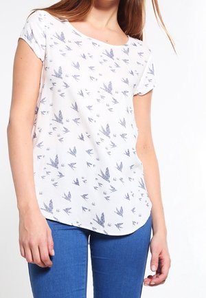 Biała bluzka z krótkim rękawem z niebieskimi nadrukami ptaków, noszona przez osobę w niebieskich dżinsach, widoczna od szyi do bioder na białym tle.