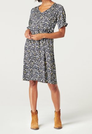 Femme portant une robe noire à fleurs mi-longue avec des manches courtes bouffantes et des bottines marron, debout les mains jointes.