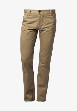 Homme marchant en pantalon beige, pull noir et orange, baskets noires, portant un sac de voyage en cuir beige et marron dans la main droite.