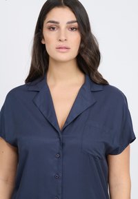 Blusa corta blu scuro a maniche corte con colletto a punta, dotata di chiusura a button front e un'unica tasca sul petto. Tessuto morbido e leggero.