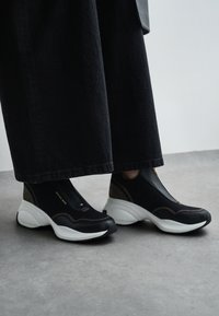 Sorte slip-ons med netoverdel og teksturerte detaljer, med en kraftig hvit såle og subtil merking på siden.