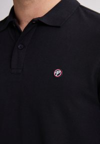 Polo shirt nera con colletto, patta con due bottoni e un piccolo logo rotondo con dettagli rossi e blu sul lato sinistro del petto. Tessuto testurizzato.