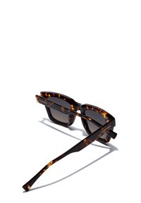Ochelari de soare cu rame tip scoică țestoasă, design pătrat, lentile brune cu gradient și brațe elegante. Include numele brandului „HAWKERS” pe lateral.
