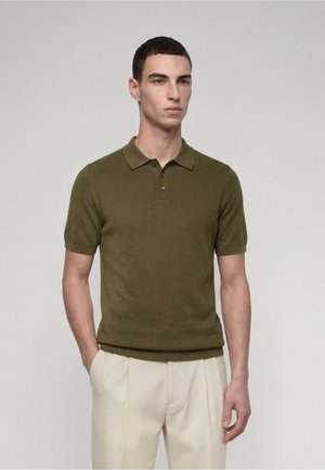 Man draagt een olijfgroen gebreid polohemd met korte mouwen en lichtbeige broek, staat met één hand in de zak tegen een effen achtergrond.