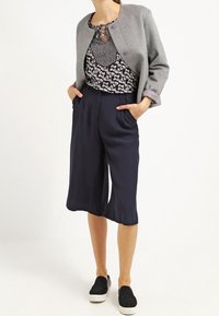 Chaqueta gris corta, blusa con estampado floral, culottes azul marino y zapatillas negras sin cordones. Diseño elegante y moderno con un ajuste relajado.