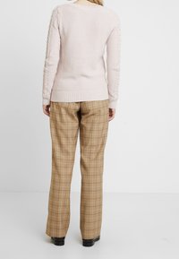 Ljusrosa stickad tröja med texturerad kabeldetaljering på ärmarna, i kombination med beige rutig byxa med rak passform och subtil glans.