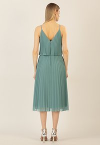 Faltenrock, t�ürkisfarbenes Kleid mit V-Ausschnitt und dünnen Trägern. Der Stoff ist leicht und fließt bis zum knielangen Saum. Silberne Absätze runden den Look ab.