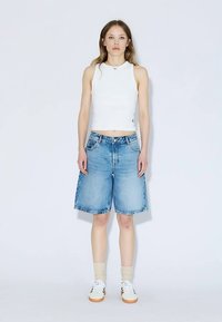 White sleeveless crop top paired with light blue denim bermuda shorts. Model wears white sneakers and beige socks. Simple, casual design.