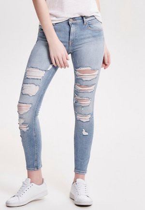 Femme portant un jean skinny bleu clair déchiré et des baskets blanches, debout avec une main près de la hanche sur un fond uni.