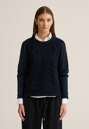 Kvinde med glat brunt hår iført en marineblå sweater med blonde mønster over en hvid skjorte med krave og sorte bukser, stående foran en beige baggrund.