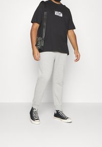 Svart kortärmad t-shirt med vit logotryck, grå sweatpants och svarta högstövlar med vita sulor. Väska slängd över kroppen.