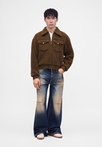 Jeune homme portant une veste sherpa marron zippée, un jean large déchiré avec des taches décolorées, et des baskets blanches, debout avec une main dans la poche.