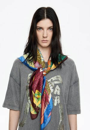 Pañuelo de seda con vibrantes patrones de pájaros y hojas en azul, amarillo, rojo y verde, atado al cuello sobre una camiseta grande gris.