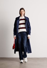 Palton de marină peste un pulover cu dungi albe și roșii, asortat cu blugi negri și adidași albi. O poșetă roșie este ținută într-o mână.