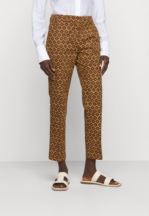 Persona que lleva pantalones ajustados amarillos con estampado, camisa blanca abotonada y sandalias blancas de punta abierta, de pie contra un fondo liso.