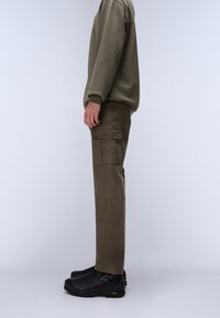 Olivegrønne cargo bukser med sidelommer, tætsiddende pasform og tykkere stof. Kombineret med sorte slip-on sko, alt sammen mod en ensfarvet baggrund.