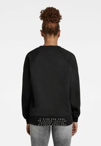 Černá mikina se žebrovaným výstřihem a dlouhými rukávy. Na zadní straně se nachází obdélníková visačka a grafický text poblíž lemu.