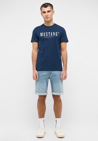 Granatowa bawełniana koszulka z białym logo "MUSTANG", w połączeniu z jasnymi szortami dżinsowymi. Model nosi białe skarpetki i kremowe sneakersy.