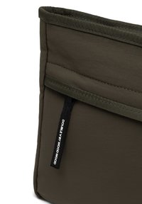 Geantă de pânză khaki cu un design matlasat și o margine superioară. Are un etichetă cu brand pe lateral și o textură netedă.