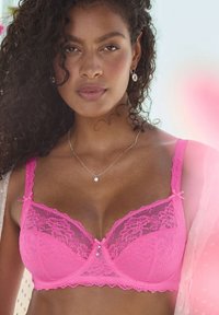 Pink lace bra with underwire, featuring floral patterns, adjustable straps, and decorative pearl and bow accents at the center.
