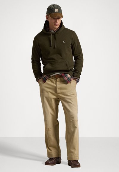 Un sweat à capuche vert olive avec une poche avant ; superposé à une chemise à carreaux rouge et noir. Pantalons beige à coupe loose avec des signes d'usure visibles ; chaussures marron.