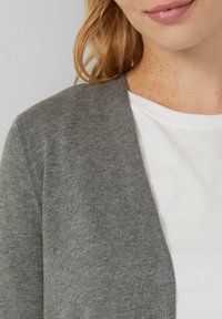 Grijze gebreide cardigan met open voorkant, lichte textuur en een subtiele heatherpatroon, gelaagd over een wit shirt met een ronde halslijn.