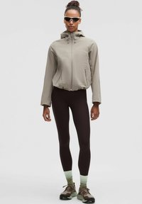 Jachetă khaki cu glugă și tiv elastic, asortată cu colanți maro închis și șosete cu accente verzi. Sneakers maro rezistenți completează ținuta.
