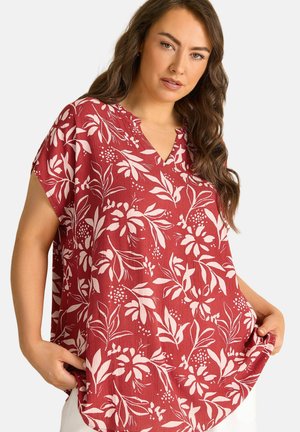 Kvinde iført løs rød bluse med hvide bladmønstre, som holder i nederste kant, hvide bukser synlige, foran ensartet lyst baggrund.